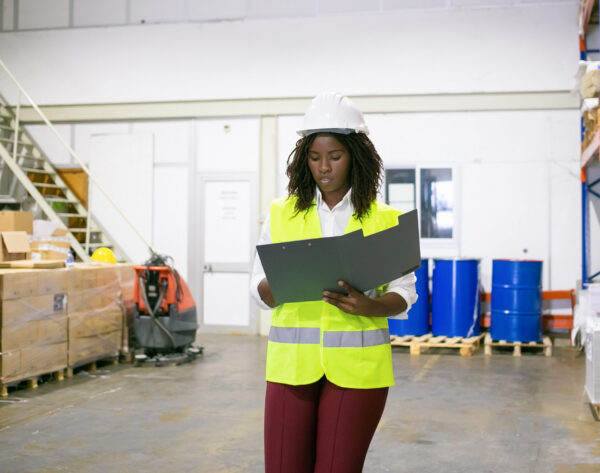 Focused female logistic employee in hardhat and safety vest walking in warehouse, carrying open folder, looking through document. Copy space, front view. Labor and inspection concept